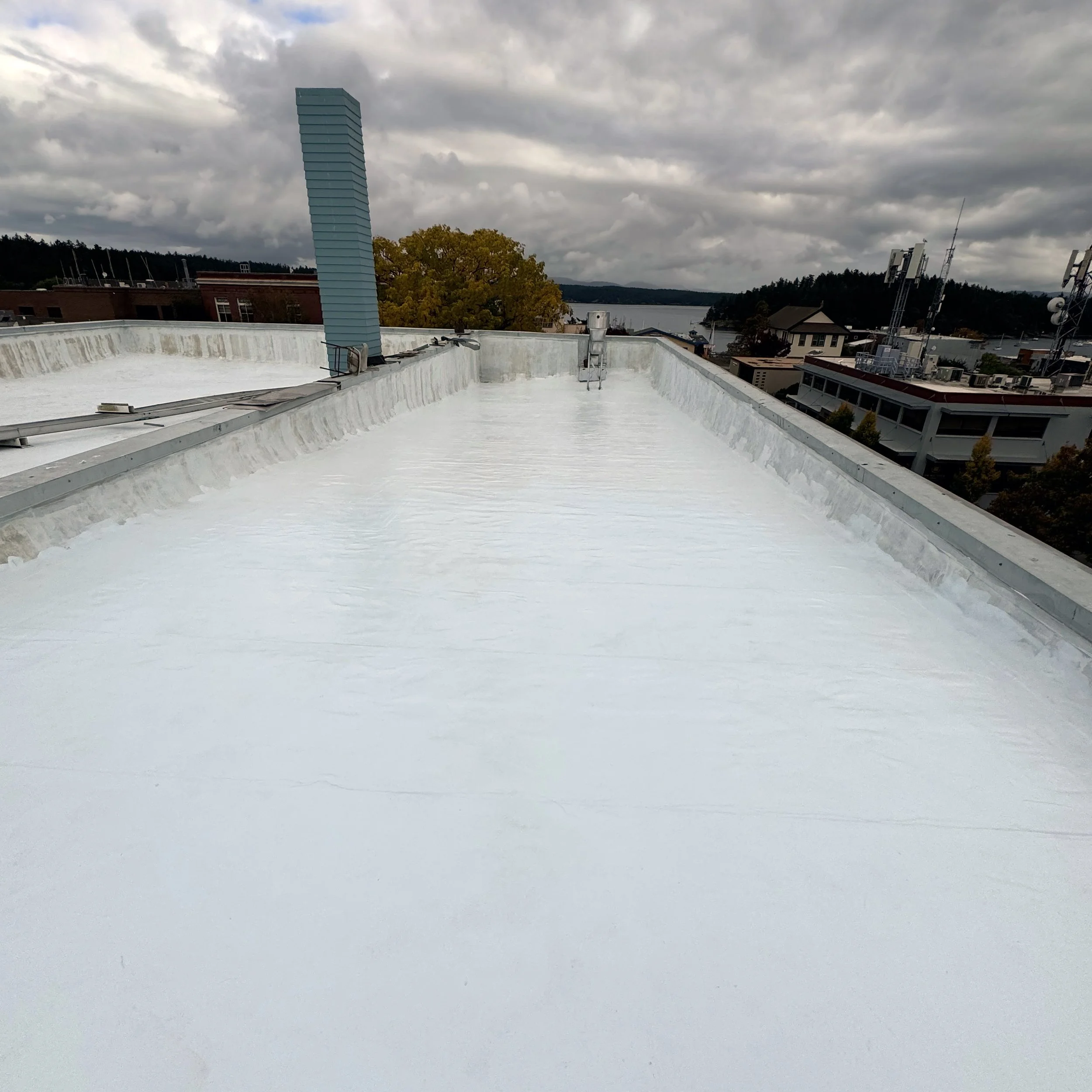 White commercial flat roof with protective coating beneath a cloudy sky overlooking nearby buildings.