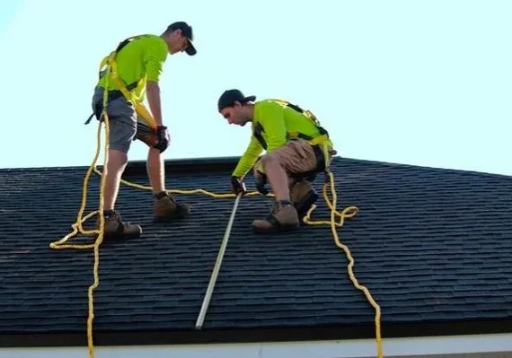Two roofing contractors measuring and aligning shingles while secured with safety harnesses
