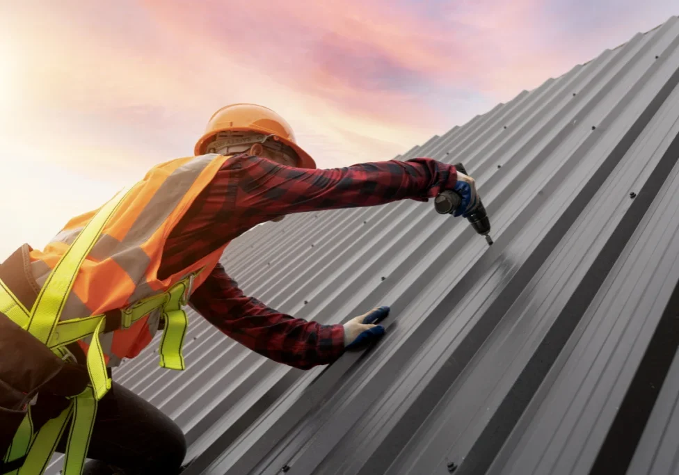 Roofing contractor installing a metal roof panel with a power drill while wearing safety gear